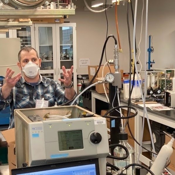 Adam is seen standing in the laboratory explaining his research. His hands are gesturing wildly. In front of him is a scanning mobility particle sizer.